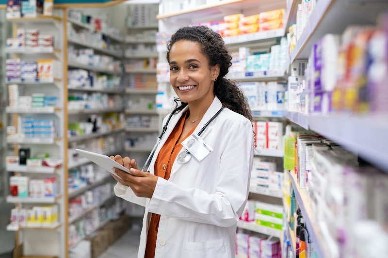 Pharmacist in a white coat at a pharmacy smiling and holding a clipboard