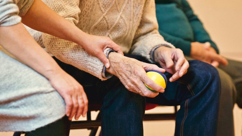 Elderly man in a waiting room with a nurse or caretaker holding his hand.